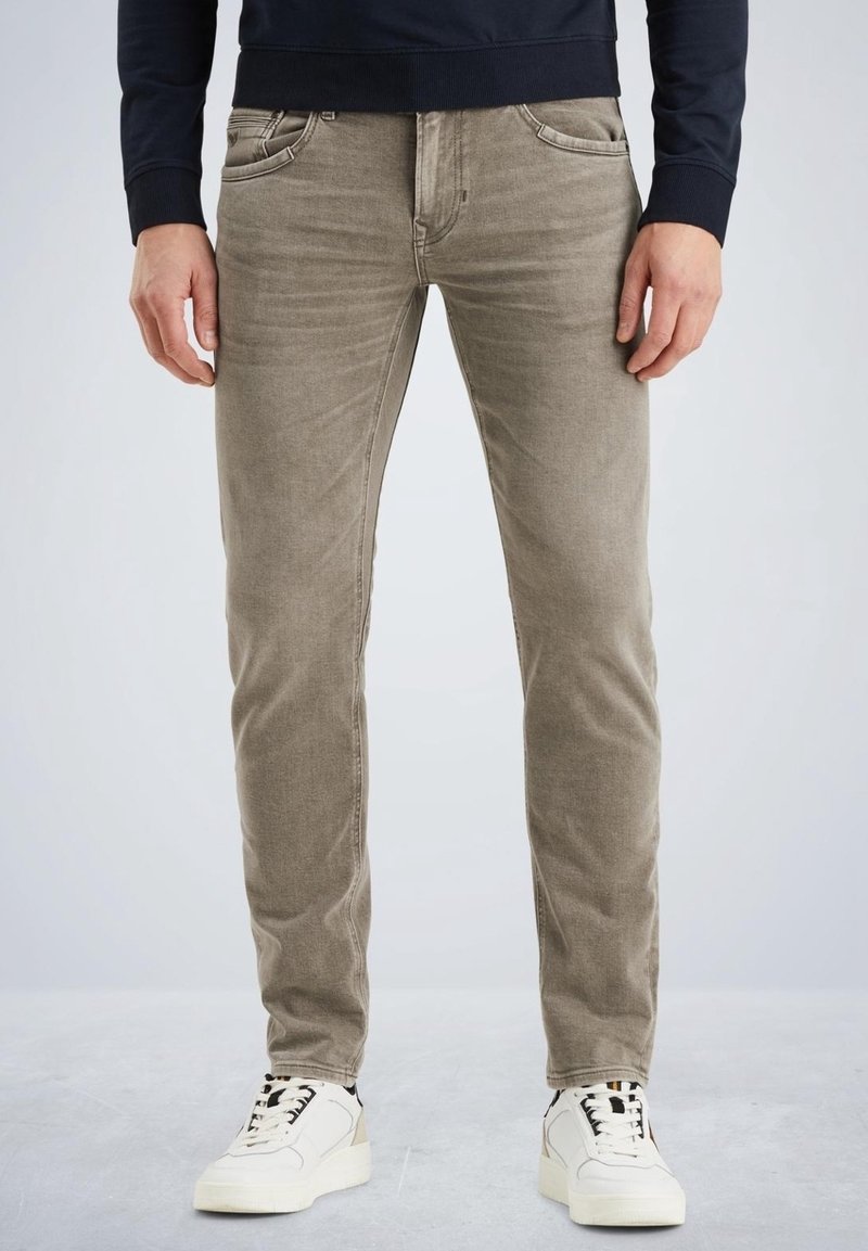Homme portant un jean beige slim-fit et des baskets blanches, debout les mains relâchées le long du corps sur un fond uni.