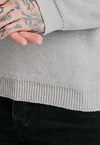 Händer med tatueringar hopknutna över en grå stickad tröja och svarta byxor, som visar detaljerad hudkonst och stoffets textur i närbild.