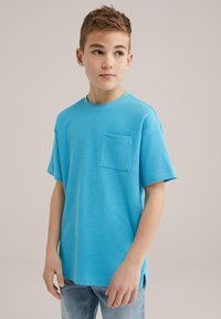 Svetlo modra majica s kratkimi rokavi iz teksturirane tkanine z okroglim izrezom in sprednjim žepom. Rob ima stranske razpoke za večje udobje.