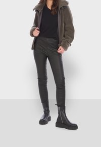 Femme portant une veste en polaire marron, un t-shirt noir à col en V, un pantalon en cuir noir et des bottines noires à fermeture éclair.