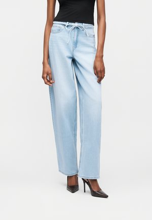 Άτομο που φοράει ανοιχτό μπλε ψηλόμεσα τζιν παντελόνια με φαρδιά πόδια και κορδόνι στη μέση, καθώς και μαύρα ψηλοτάκουνα παπούτσια με μυτερή μύτη, στέκεται μπροστά σε λευκό φόντο.