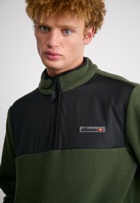 Green fleece pullover with a black fabric upper section, high collar, and zipper. Features a small logo patch on the chest. Textured surface.