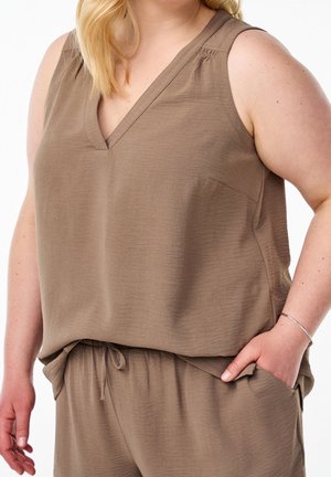 Vrouw draagt mouwloze bruine V-hals top en bijpassende broek met taillekoord, hand in de zak, tegen witte achtergrond.