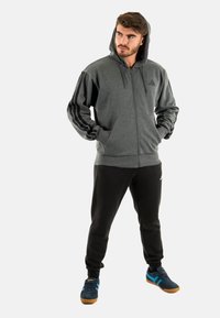 Felpa grigia con zip e cappuccio, dotata di strisce nere decorative sulle maniche, abbinata a pantaloni della tuta neri e scarpe da ginnastica blu.