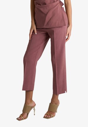 Donna che indossa pantaloni malva corti e affusolati con spacchi laterali abbinati a sandali beige e oro con tacco alto, in piedi su sfondo bianco.