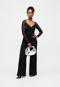 Femme portant une combinaison noire à jambes larges avec des manches longues transparentes et des talons noirs, tenant un sac à main métallisé argenté sur un fond blanc.