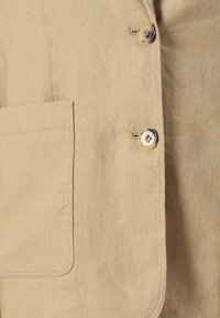 Blazer beige in lino con una tasca sul petto, bottoni argentati e finitura in tessuto testurizzato. Forma sartoriale con linee pulite.