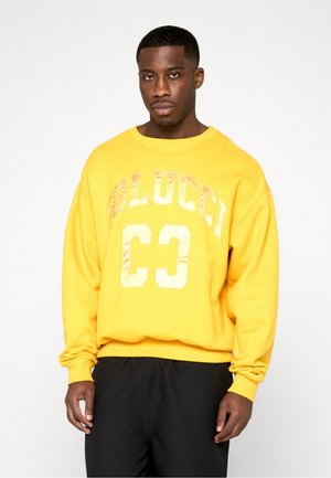 Homme portant un sweat-shirt jaune avec "COLUCCI 63" en lettres brillantes, debout devant un fond blanc uni.