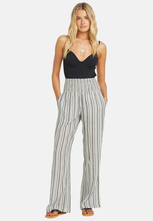 Vrouw met lang blond haar, gekleed in een zwart hemdje, hoog uitgesneden wijde gestreepte broek en sandalen, met haar handen in haar zakken.