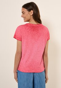 T-shirt a maniche corte e collo rotondo di un rosa vivace, con una finitura morbida e testurizzata. Indossato con pantaloni larghi di un blu chiaro, visto da dietro.