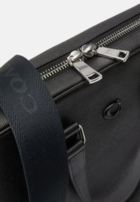 Crna kožna torba s teksturiranim finišem, sadrži srebrne patent zatvarače, tamnu platnenu traku i suptilnu oznaku.
