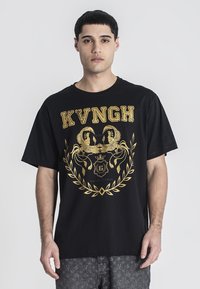 Camiseta de algodón negra con un gran gráfico dorado que presenta un motivo de fénix, el texto "KVNGH" y un diseño de laurel. Cuello redondo, mangas cortas.