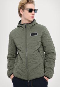 Olive green quilted jacket with a hood, zippered front, logo patch on chest, elastic cuffs, and side pockets. Smooth texture, lightweight design.