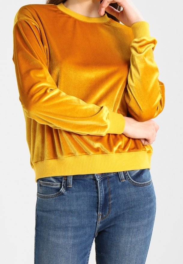 Sudadera de terciopelo amarillo con mangas largas y escote redondeado, combinada con jeans de mezclilla azul. Textura suave y corte relajado.