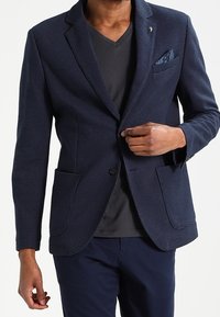 Granatowa marynarka o fakturowanej powierzchni, z dwoma przednimi kieszeniami i poszetką. Lekka tkanina, dopasowany krój, bez widocznych zapięć.