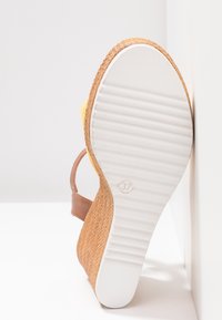 Sandale compensée avec talon en corde texturée couleur tan, semelle blanche rainurée indiquant la taille 37, et bride de cheville marron appuyée contre une surface blanche.