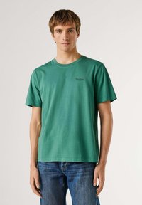 Mladý muž v obyčajnom zelenom tričku s krátkym rukávom a modrých džínsoch stojaci pred svetlým neutrálnym pozadím.