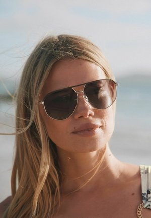Jeune femme aux longs cheveux blonds portant des lunettes de soleil aviateur foncées, en extérieur avec un arrière-plan flou de plage par une journée ensoleillée.