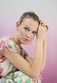 Blumenmusterkleid aus transparenter Stoff in Pastellfarben; hellgrün, pink und weiß. Modell sitzt, hält leicht die Hände zusammen, vor einem Farbverlauf-Hintergrund in Pink.