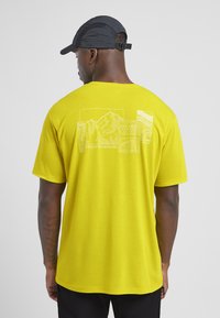 T-shirt giallo a maniche corte con una grafica di montagna e motivi geometrici bianchi sul retro. Tessuto morbido, vestibilità comoda.