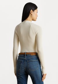 Vrouw met lang zwart haar draagt een crèmekleurig ribgebreid shirt met lange mouwen, blauwe jeans en een bruine riem, staande met een lichte zijwaartse draai.