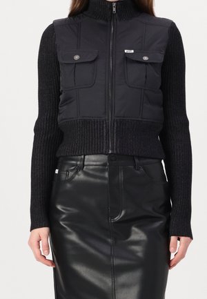 Black zip-up cropped jacket with knit sleeves and two flap pockets paired with a high-waisted black leather skirt.