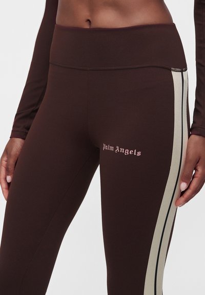 Κοντινό πλάνο ενός ατόμου που φοράει καφέ leggings με μπεζ και μαύρες πλαϊνές ρίγες και ροζ λογότυπο "Palm Angels" στο αριστερό μηρό.