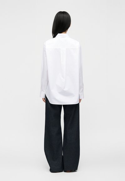 Femme aux longs cheveux noirs portant une chemise blanche ample boutonnée et un pantalon noir à jambes larges, debout de dos sur un fond uni.