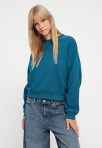 Sudadera recortada color teal con cuello redondo y hombros caídos, combinada con jeans de corte relajado en azul claro, que cuentan con bolsillos y un botón.