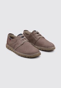 Zapatos sintéticos marrones con una textura suave, diseño de cordones y paneles perforados. La suela presenta un patrón moteado en contraste.
