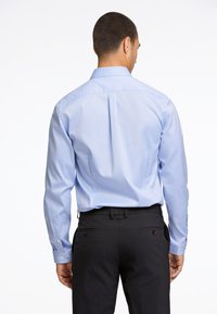 Camicia da uomo azzurra a maniche lunghe con polsini abbottonati, dettaglio sullo scollo posteriore e design infilato, abbinata a pantaloni scuri sartoriali.