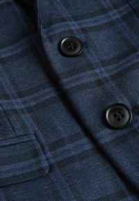 Tessuto a quadri blu navy, texture liscia, con due bottoni neri e una tasca. Il motivo include linee blu sottili e scure.