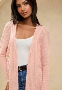 Világos rózsaszín kötött kardigán texturált mintákkal, nyitott elülső részével és hosszú ujjával. Fehér ujjatlan topon viselve, farmernadrággal párosítva.