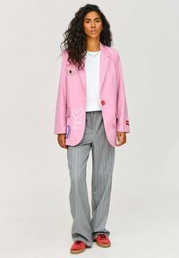 Růžový blazer s grafikou oka a srdce, se singledem červeným knoflíkem, kombinovaný se šedými pruhovanými kalhotami a červenými teniskami, nošený přes bílou košili.