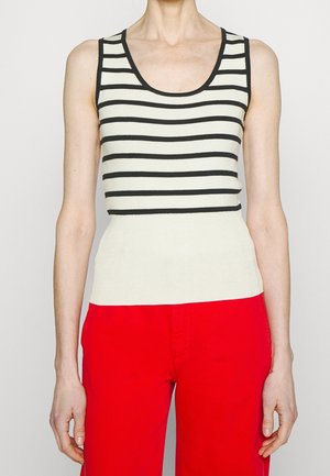 Mujer con top blanco sin mangas con rayas horizontales negras y pantalones rojos brillantes, de pie contra un fondo liso.