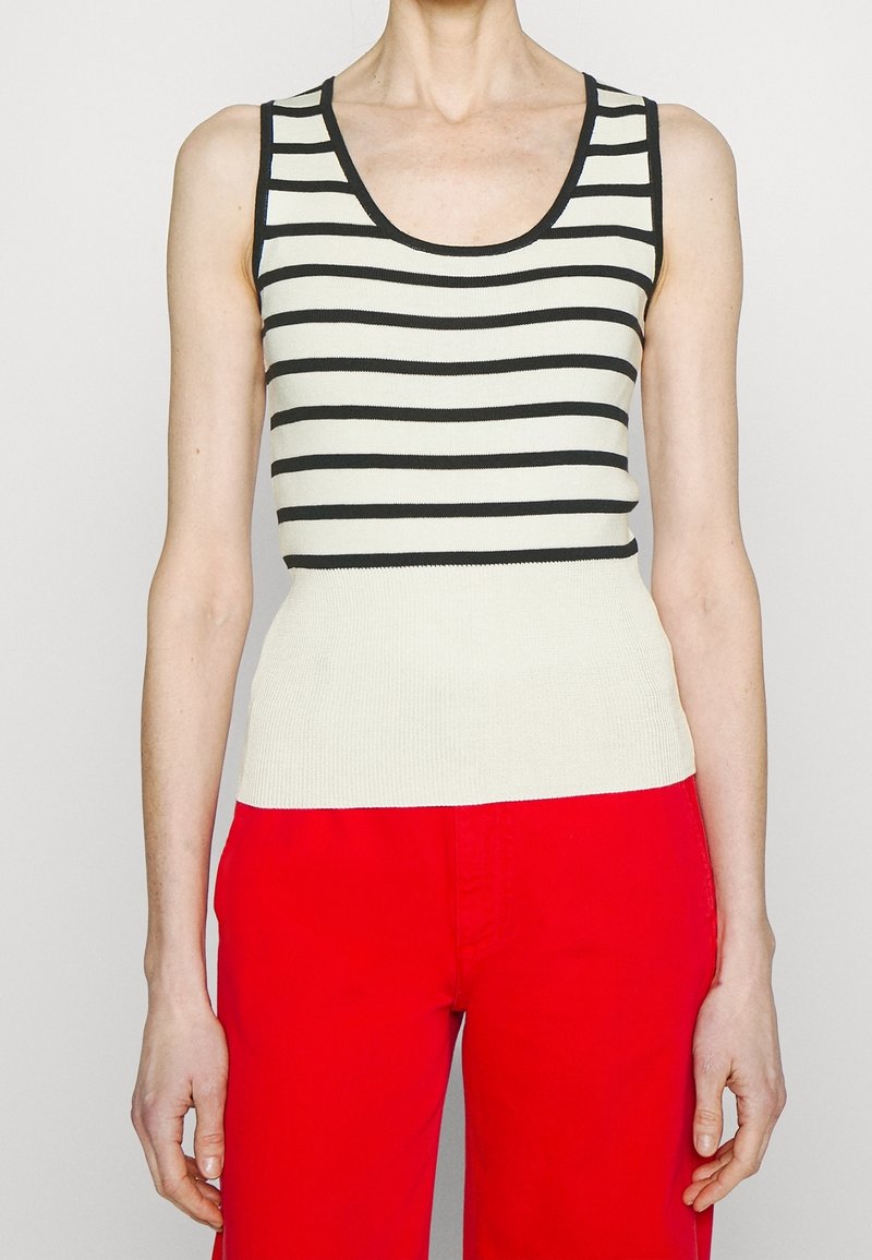 Femme portant un haut blanc sans manches à rayures horizontales noires et un pantalon rouge vif, debout devant un fond uni.