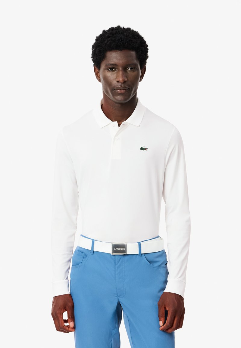 Vit långärmad polo med en grön krokodil-logotyp; matchad med blå byxor och ett vitt bälte med en silvrig spänne.