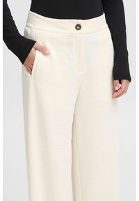 Witte, gestructureerde broek met een wijde pijp, voorzien van zijzakken en een sluiting met een knoop aan de voorzijde. De stof heeft een subtiel patroon voor extra detail.