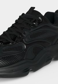 Baskets épaisses noires avec panneaux en mesh mixte et textures brillantes, semelle épaisse et lacets noirs, vues de côté en angle.