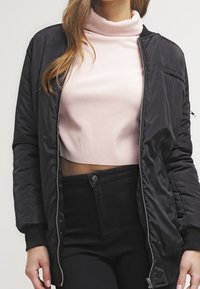 Schwarze Bomberjacke mit glatter Textur, Reißverschluss vorn und geripptem Kragen, getragen über einem pinken gerippten Rollkragenpullover und hochtaillierten schwarzen Hosen.