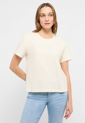 Frau mit hellbraunen Haaren, die ein cremefarbenes kurzärmliges Hemd und hellblaue Jeans trägt, steht mit einem Arm hinter dem Rücken.