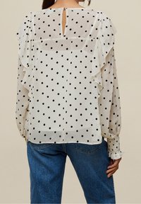 Weißes Blusenshirt mit schwarzen Polka-Dots, Rüschensäumen und einem Schlüssellochdetail am Rücken. Der Stoff ist sheer mit einer glatten Textur. Kombiniert mit Jeans.