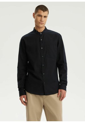 Młody mężczyzna ubrany w czarną koszulę z mandaryńskim kołnierzykiem i beżowe spodnie, stojący na jednolitym jasnym tle.