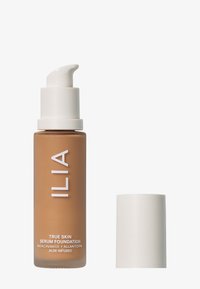 True Skin Serum Foundation i en frostet brun flaske med en hvit pumpe. Etiketten inneholder produktdetaljer og nøkkelingredienser. Lokket er hvitt.