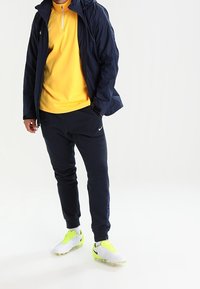 Marynarka w kolorze granatowym, żółta bluza na zamek, granatowe joggery i białe buty sportowe z neonowymi akcentami. Sportowy design, lekkie materiały i dopasowany krój.