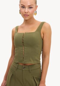 Femme portant un crop top sans manches vert olive avec fermetures à crochets sur le devant et un pantalon taille haute assorti, face à l'avant sur fond blanc.