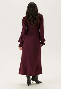 Robe longue bordeaux en tissu texturé, avec des manches bouffantes et une taille cintrée. Complétée par des bottes noires jusqu'au genou.
