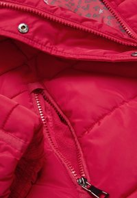 Interior de chaqueta acolchada roja, con una textura suave y un forro de patrón gris en contraste. Incluye un bolsillo con cremallera y puños elásticos.
