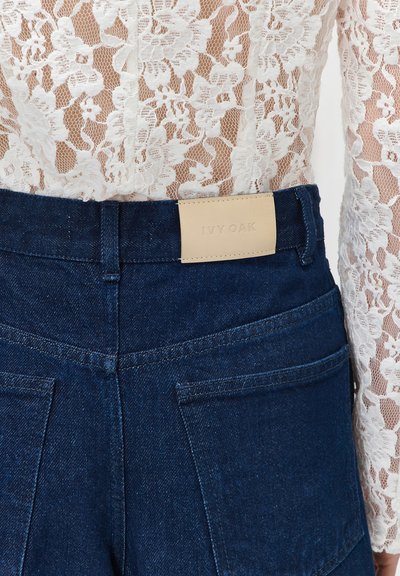 Κοντινό πλάνο στη μέση πίσω σκέλος σκούρου μπλε τζιν με ετικέτα IVY OAK σε ανοιχτό ταμπά χρώμα, συνδυασμένο με λευκό τοπ με δαντέλα και λουλούδια.