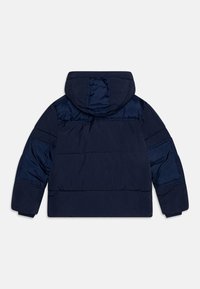 Tamno plava jakna od puffa s kapuljačom. Ima tkanje s rombovima i elastične manžete. Glatka tekstura s laganim sjajem. Prikazani pogled straga.